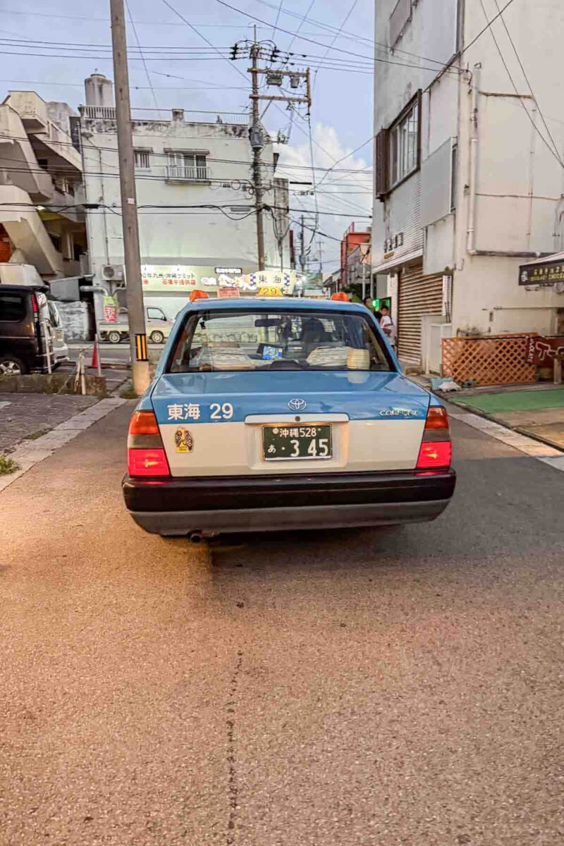 Blue and white Toyota taxi driving down a quiet street in Ishigaki, Japan at dusk with low buildings and overhead power lines. A typical local taxi option for getting around Okinawa with kids outside major cities.
