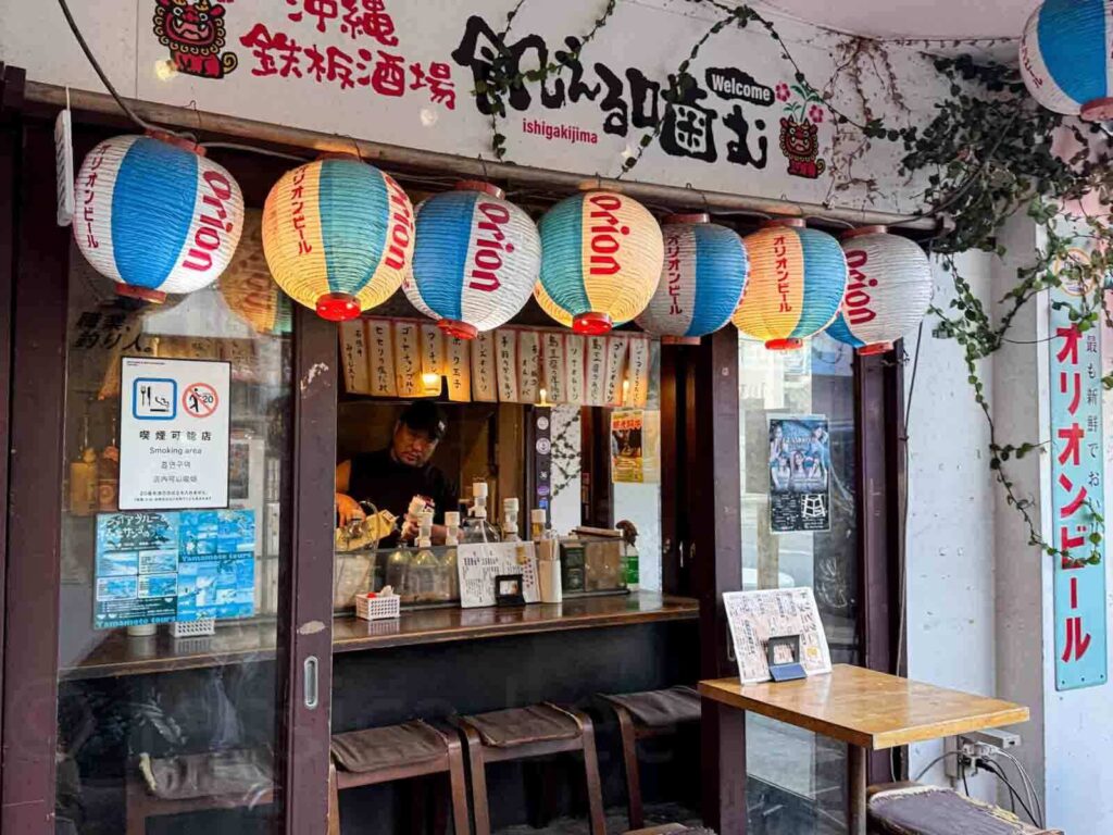 Small restaurant stall in Ishigaki with colorful white, blue and red paper lanterns labeled "Orion" hanging above the counter and a sign reading "Welcome Ishigakijima". Small local spots like this are fun for trying regional food.