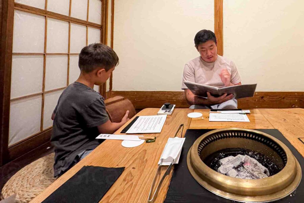 Father and child seated at a traditional low wooden table with shoji doors inside a yakiniku restaurant in Ishigaki while looking over the menu beside a charcoal grill. Unique meals like this can be memorable for kids visiting Japan.