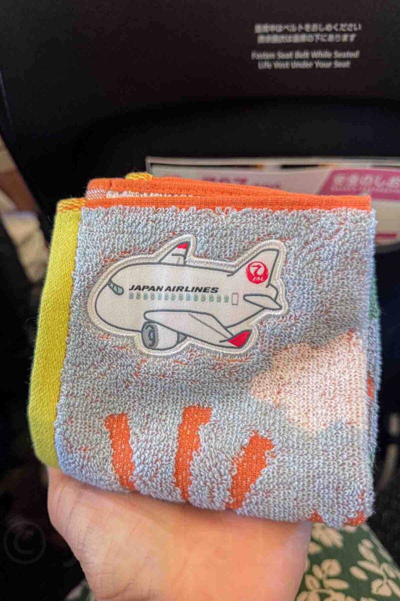 Small yellow, orange and blue hand towel with an embroidered airplane and text reading "Japan Airlines" held in a hand on a flight. A thoughtful detail that makes flying with kids in Japan more enjoyable.