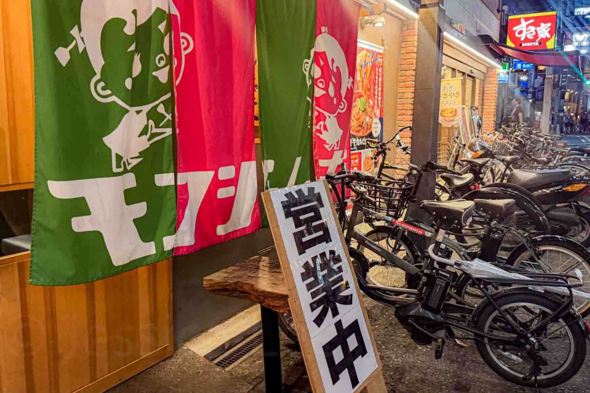 Narrow street with bicycles parked outside a small restaurant entrance covered by red and green noren curtains. A glimpse into everyday dining options in Japan.