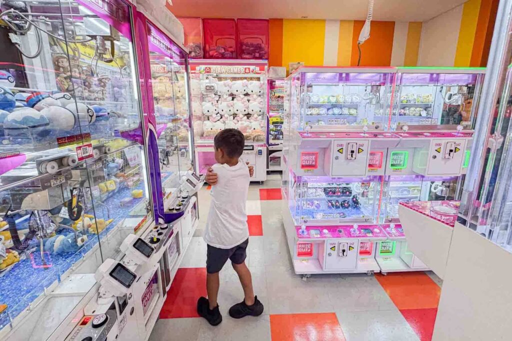 Child standing inside a Taito Station Tokyo arcade surrounded by bright white and glowing claw machines filled with plush prizes. Arcades are a common and exciting thing to do with kids in Japan.