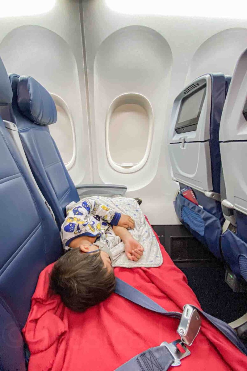 Child in pajamas sleeps stretched across airplane seats with a pillow and a red blanket during an overnight flight showing how to help kids sleep on a plane.