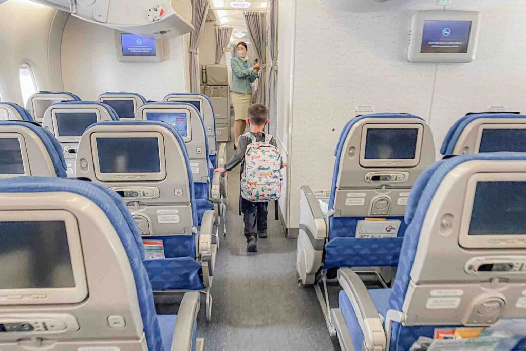 A young child wearing a patterned airplane backpack walks down the plane's aisle toward the exit while a flight attendant stands nearby. The moment shows an independent travel kid leaving the plane after a long international flight.