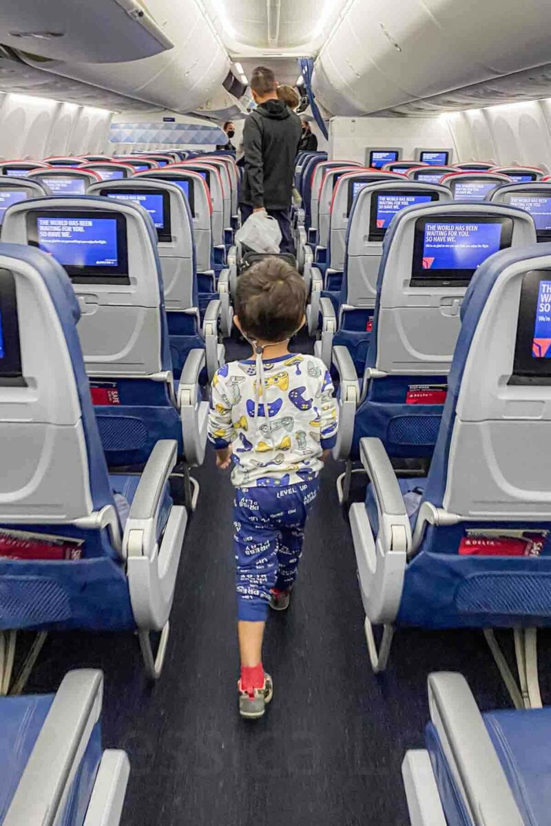 Young child in pajamas with graphic gaming controllers on them walks down the airplane aisle past screens showing "The world has been waiting for you so have we" during deboarding of a Delta fligh.