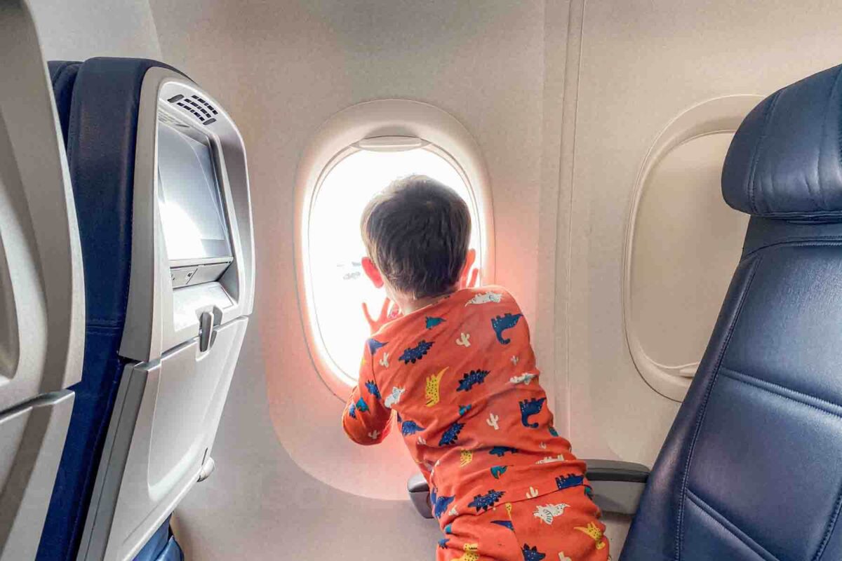 Child in orange pajamas with a dinosaur print leans toward the window looking outside during a flight.