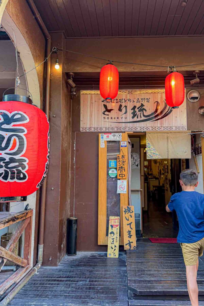 Young child walking into a small Japanese restaurant in Ishigaki with red lanterns hanging outside and wooden signs with Japanese writing by the entrance. The welcoming storefront and casual setting highlight what eating in Japan with kids can look like when exploring local eateries.