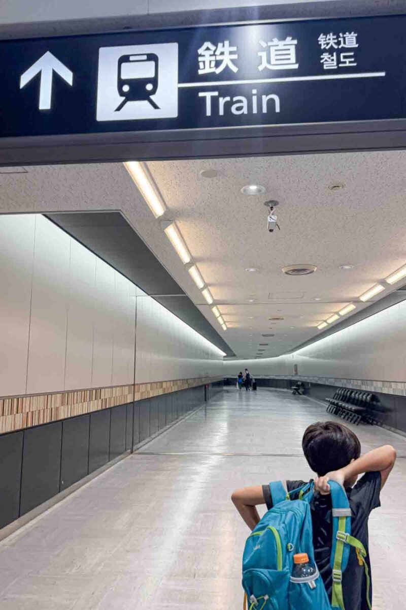 Child with a backpack walking through a station corridor under a sign that reads "Train" in English and Japanese with arrows pointing ahead. Clear signage helps simplify getting around Japan with kids in stations.