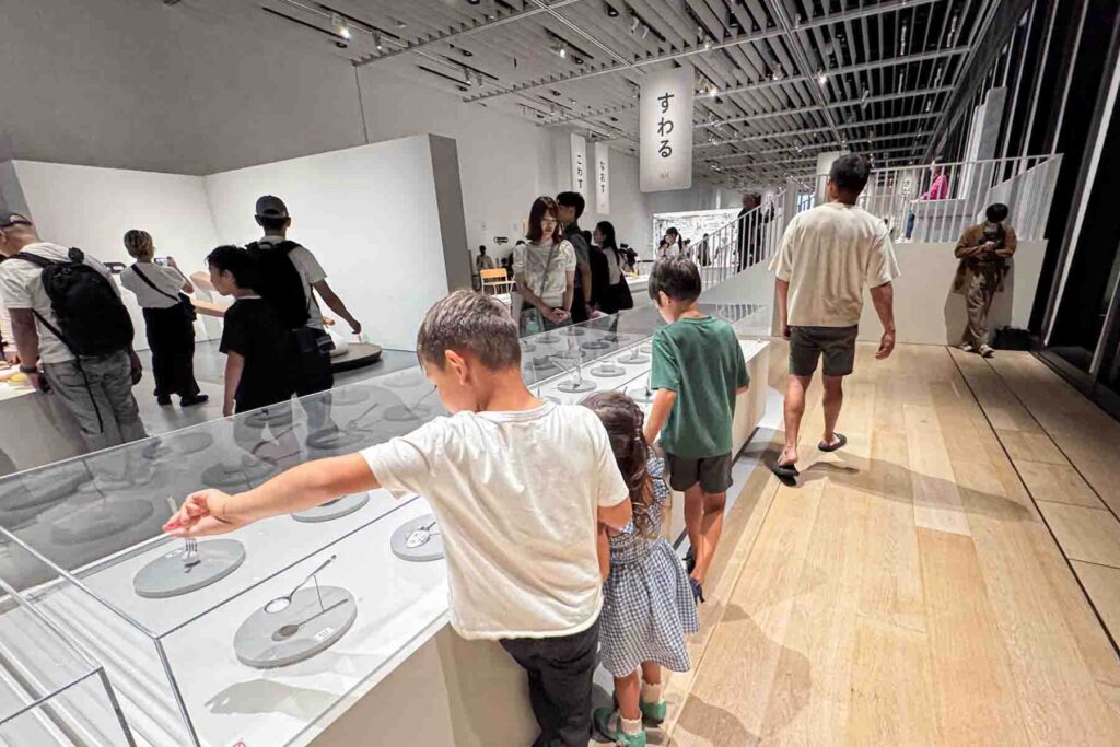 Children exploring a bright design museum exhibit at Tokyo Node with white display cases and everyday objects laid out in rows. Hands on museums like this can keep kids engaged while sightseeing.