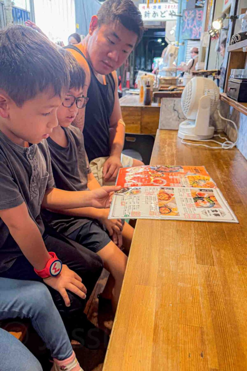 Father and two young boys sit at a small wooden counter restaurant in Osaka looking through a colorful Korean menu together. A relatable moment of dining in Japan with kids.