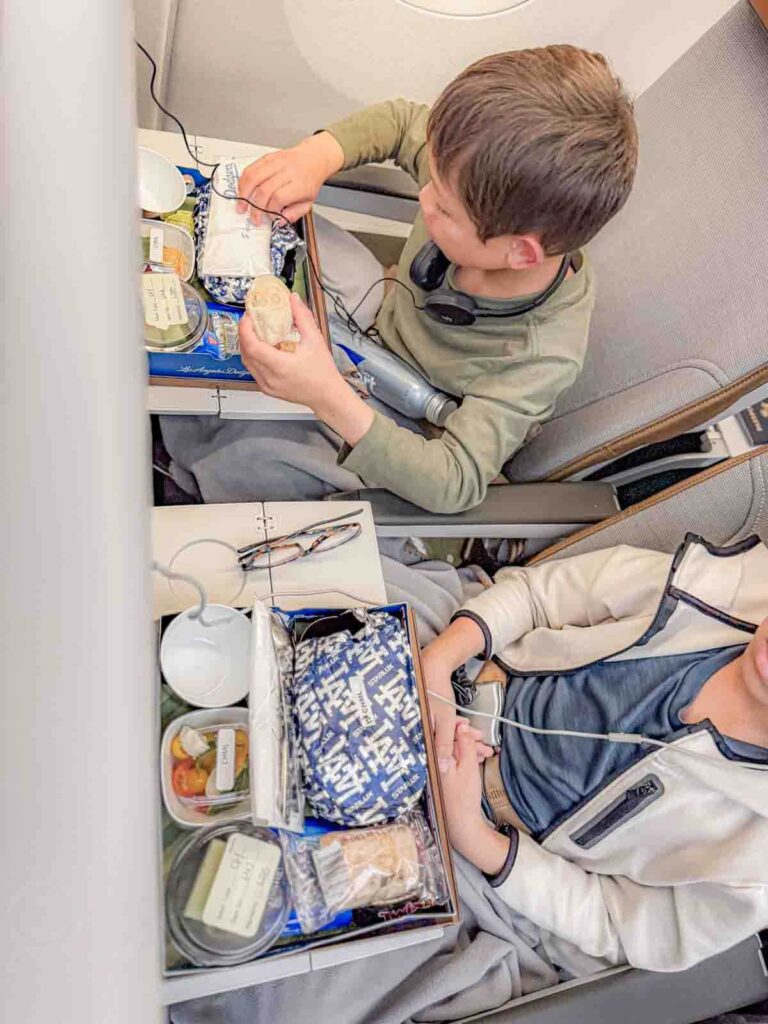 Two children sit in airplane seats on a Starlux flight eating children’s meals with trays of bread fruit and packaged food while wearing headphones.