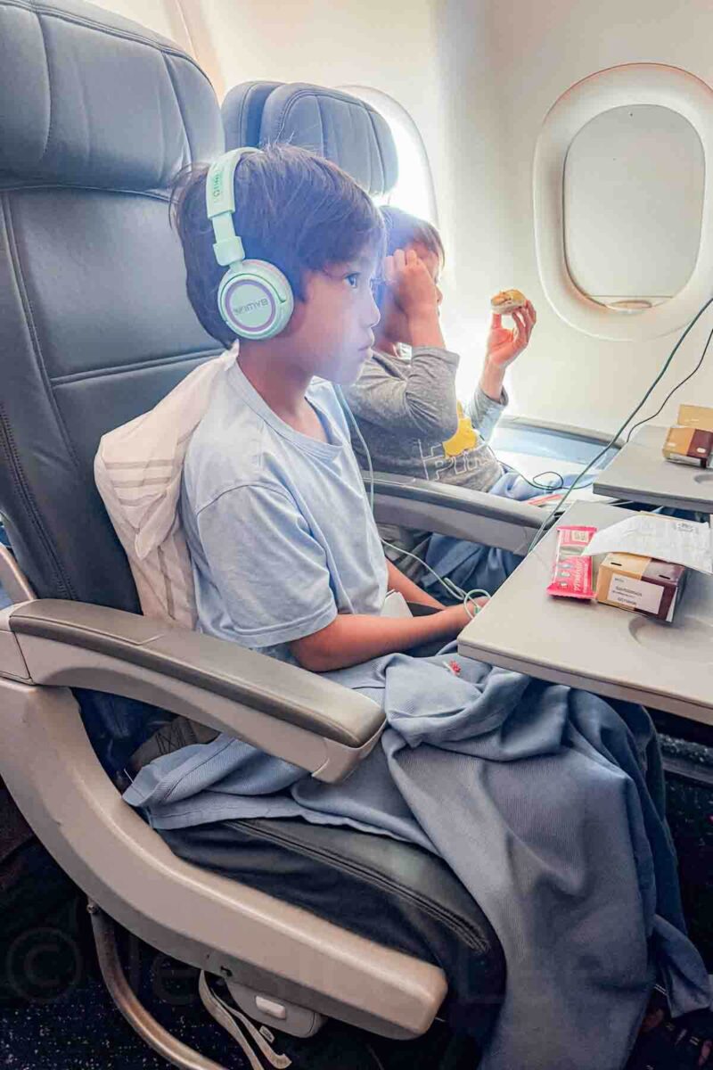 Two boys wear headphones while watching screens and eating snacks during a long flight with kids.