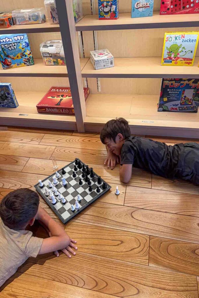 Two children lying on a wooden floor playing chess beside a shelf of games in a Mimaru hotel lounge in Osaka. Downtime activities like this help balance a busy Japan itinerary with kids.