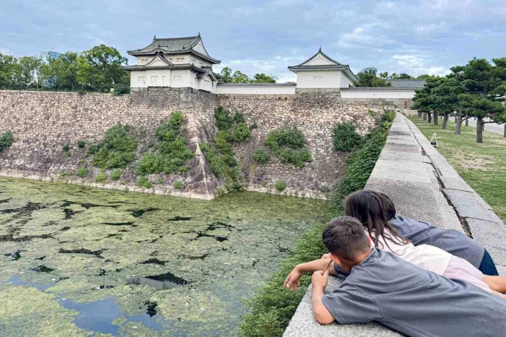 Three children laying their bellies across a wide stone wall, below is a moat filled with a bit of water and lot of moss and lily pads, the moat a barrier to the big fortress-like stone wall protecting Osaka Castle. The wide grounds give kids room to explore while sightseeing.