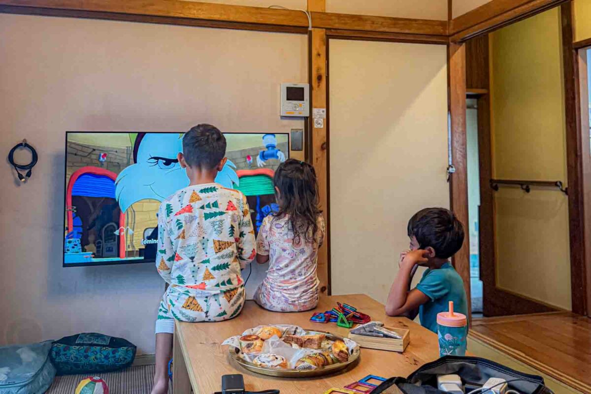 Three kids in pajamas sit at a low table watching a colorful cartoon on TV in a traditional Japanese room. Breakfast pastries and toys are scattered on the table during a relaxed morning in Japan with kids.