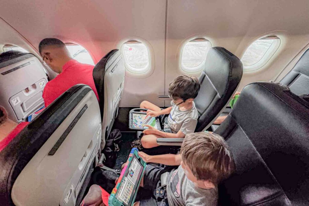 Two brothers sit in window and middle airplane seats playing games on iPads during the flight. Tablets help keep children entertained while traveling long distance.