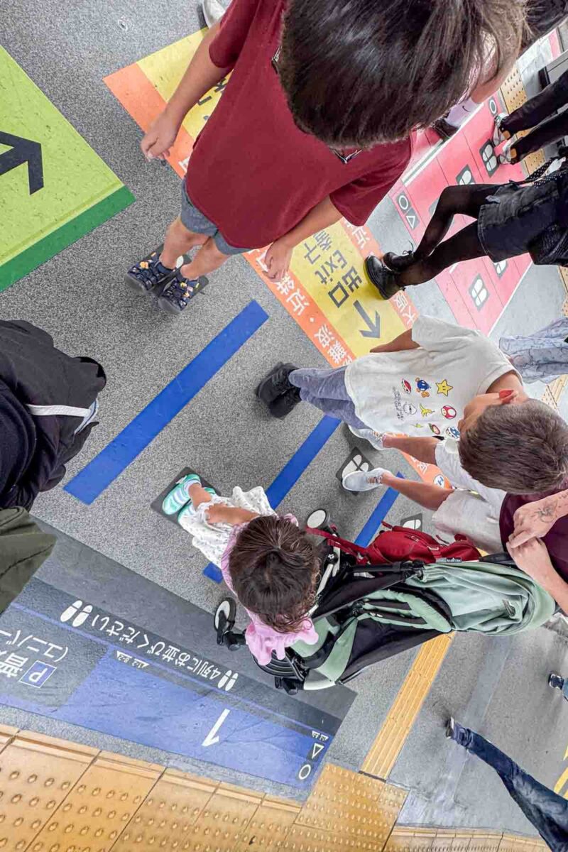 Children standing on marked platform lines with arrows and symbols showing where to queue for the train. Organized boarding areas make Japan train travel with kids more predictable and manageable.