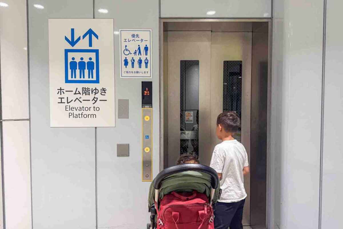 Child stands beside a stroller waiting at a station elevator next to a sign that reads "Elevator to Platform" and priority icons for accessibility. Highlights how elevators support getting around Japan with kids and strollers in train stations.