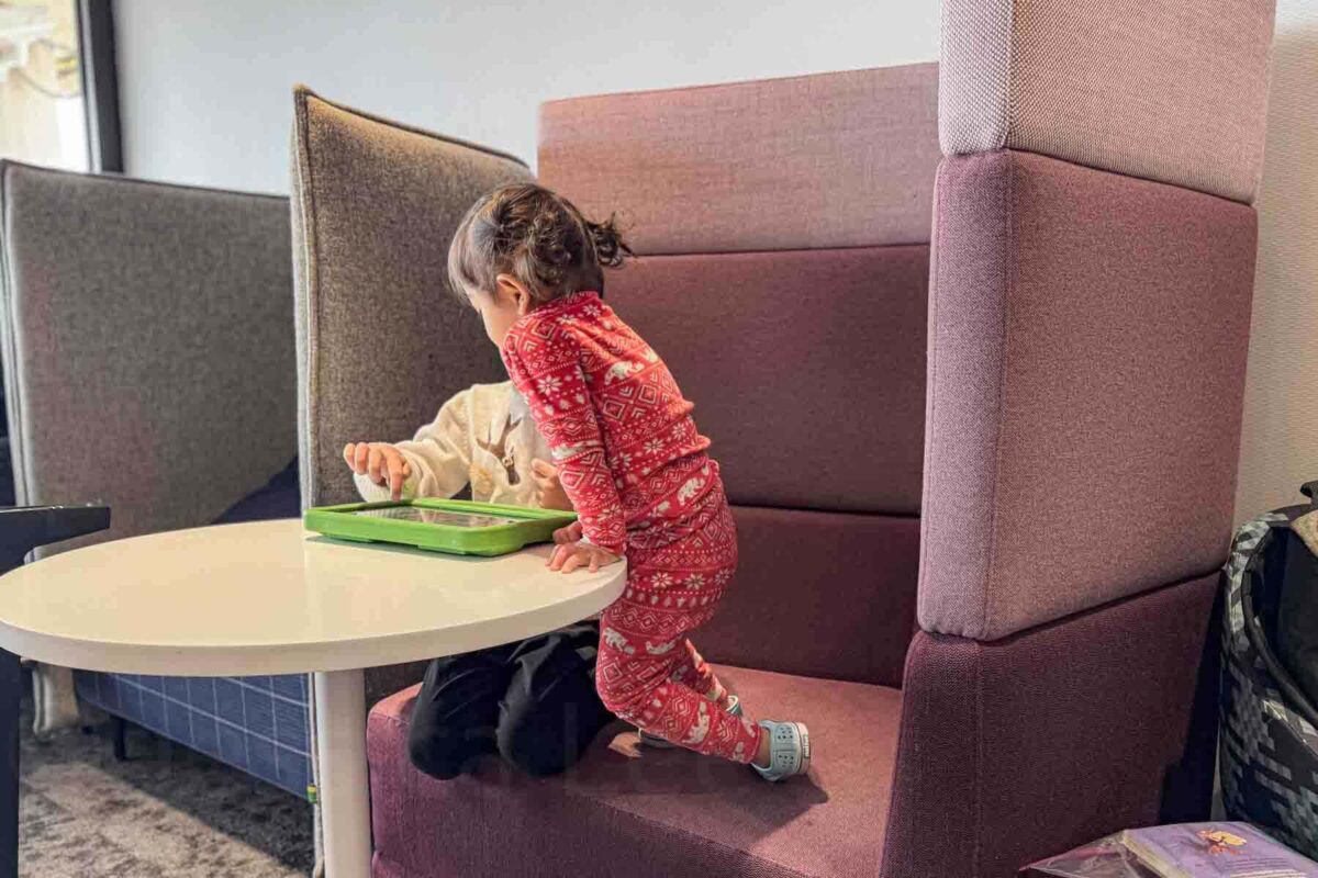 Two young children sit in an airport lounge semi-circle couch seat using a tablet together before departure.