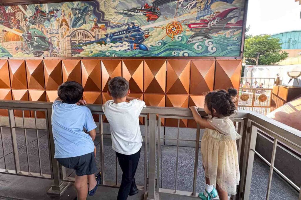 Three children leaning on a railing waiting in line for the monorail-like train at Tokyo DisneySea while looking at a colorful mural in front of them. Theme park days like this are often worth adding to a Japan family trip itinerary.