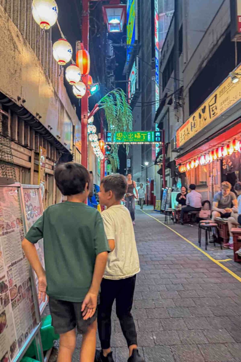 Two children walk down a narrow alley in Shinjuku lined with glowing lanterns and small restaurants with menus displayed outside the Omoide Yokocho area. Captures a lively evening street scene while exploring Tokyo with kids.