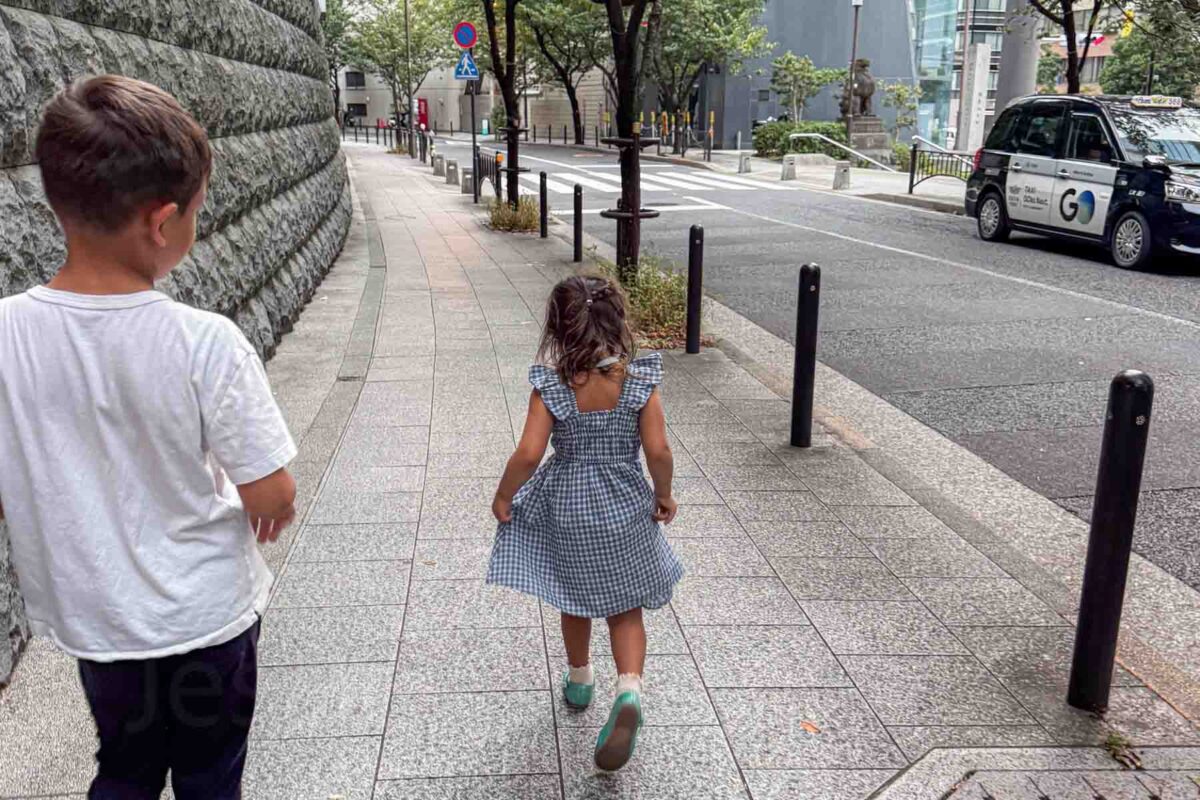 Two children walk along a clean Tokyo sidewalk beside a quiet street with a GO taxi parked nearby. Shows how walking and taxis combine easily when getting around Japan with kids.