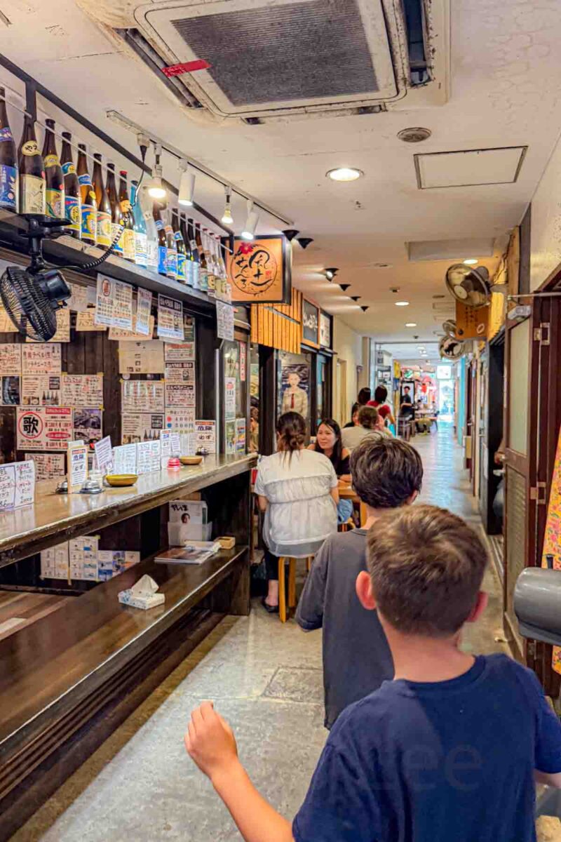 Two kids walk through a narrow indoor food market lined with small Japanese restaurants and bar seating, with menus and signs in Japanese covering the walls. The lively setting shows a casual option for eating in Japan with kids at local dining spots.