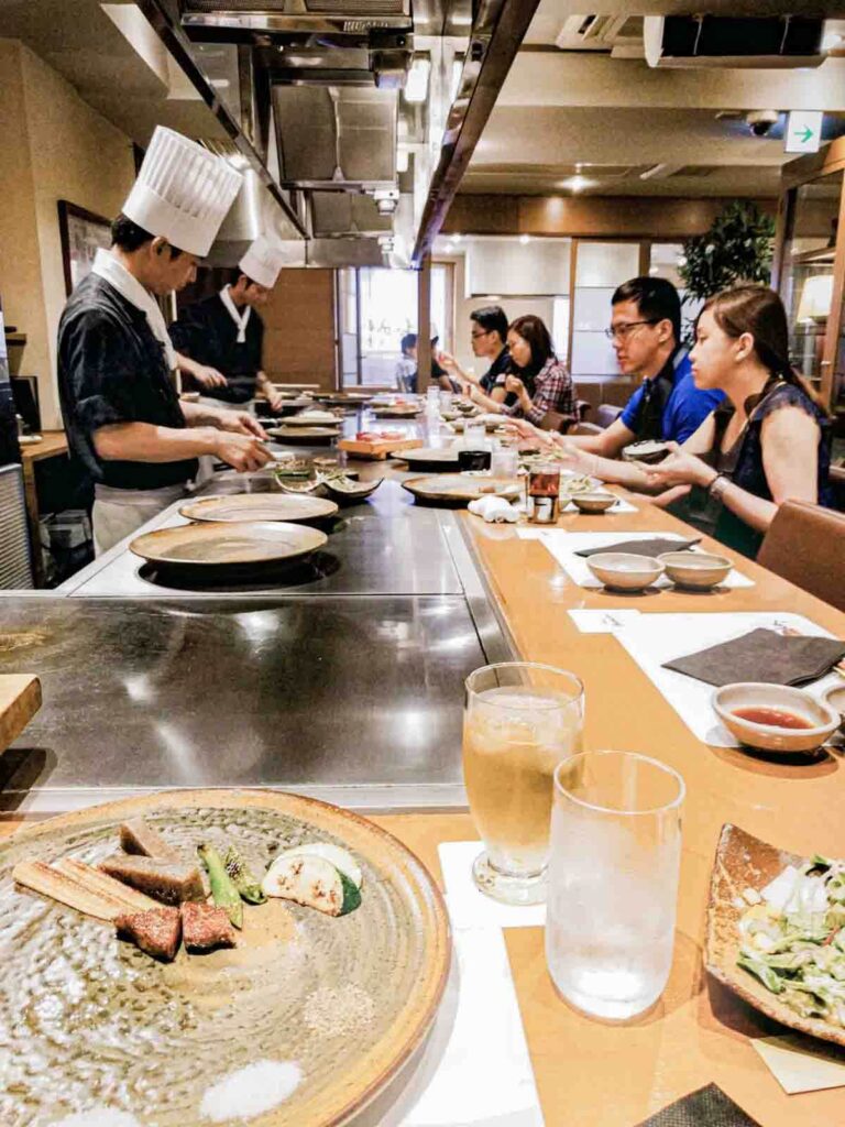 Diners seated at a kobe beef teppanyaki counter while chefs in tall white hats cook on a flat grill in front of them. Interactive teppanyaki meals like this can be especially fun for kids when traveling to Japan.
