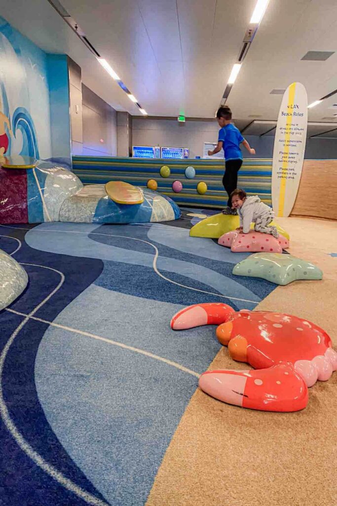Children climb on colorful beach themed soft structures including a giant red crab, colorful starfish, and a wave with a surboard in the Los Angeles international terminal airport play area. A sign nearby reads "LAX Beach Rules".