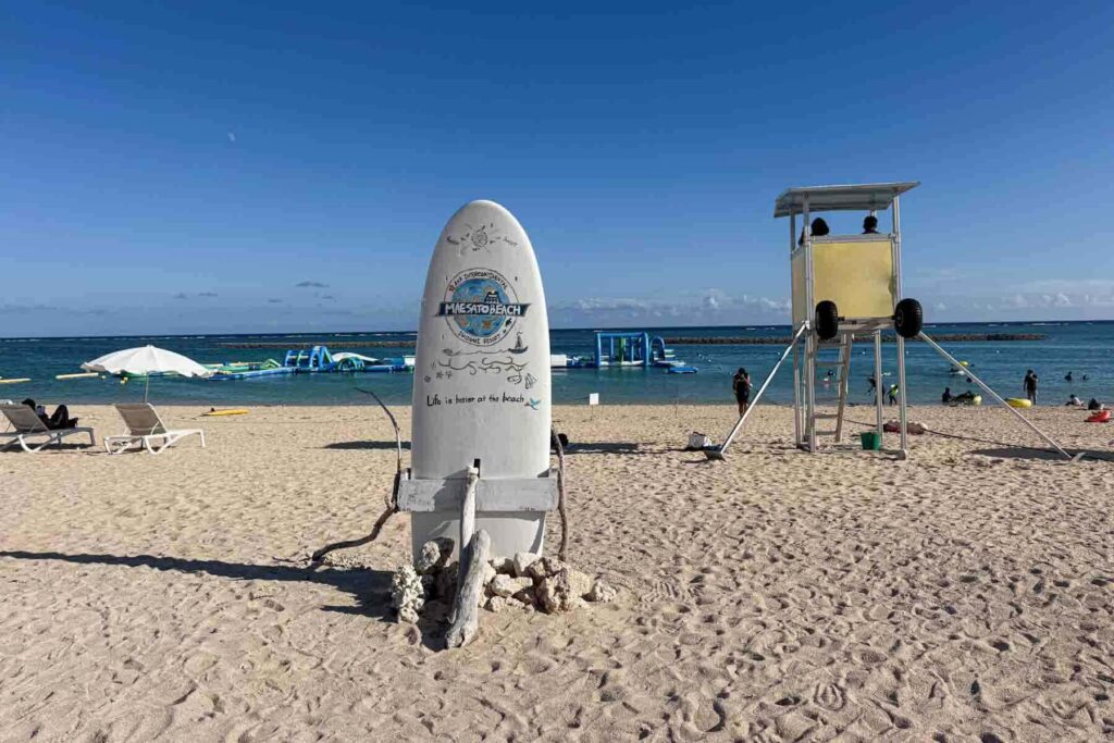Beach scene at Maesato Beach in Ishigaki with a white upright surfboard sign that reads "MAESATO BEACH" and "Life is better at the beach". A lifeguard tower stands beside the water, and an inflatable obstacle course floats in the water.