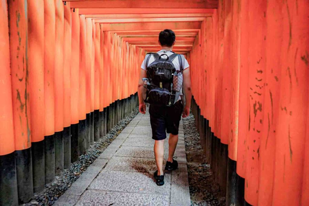 Traveler walking through tight rows of bright red torii gates at Fushimi Inari Shrine in Kyoto. Famous places like this often become highlights of a first trip to Japan.