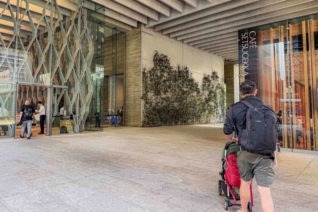 Father wearing a black backpack pushing a yoyo stroller with a red backpack on it past a modern building with a green wall in Ginza. They’re approaching a modern glass see-through cafe. Walkable districts like Ginza are a convenient option for a Tokyo trip.