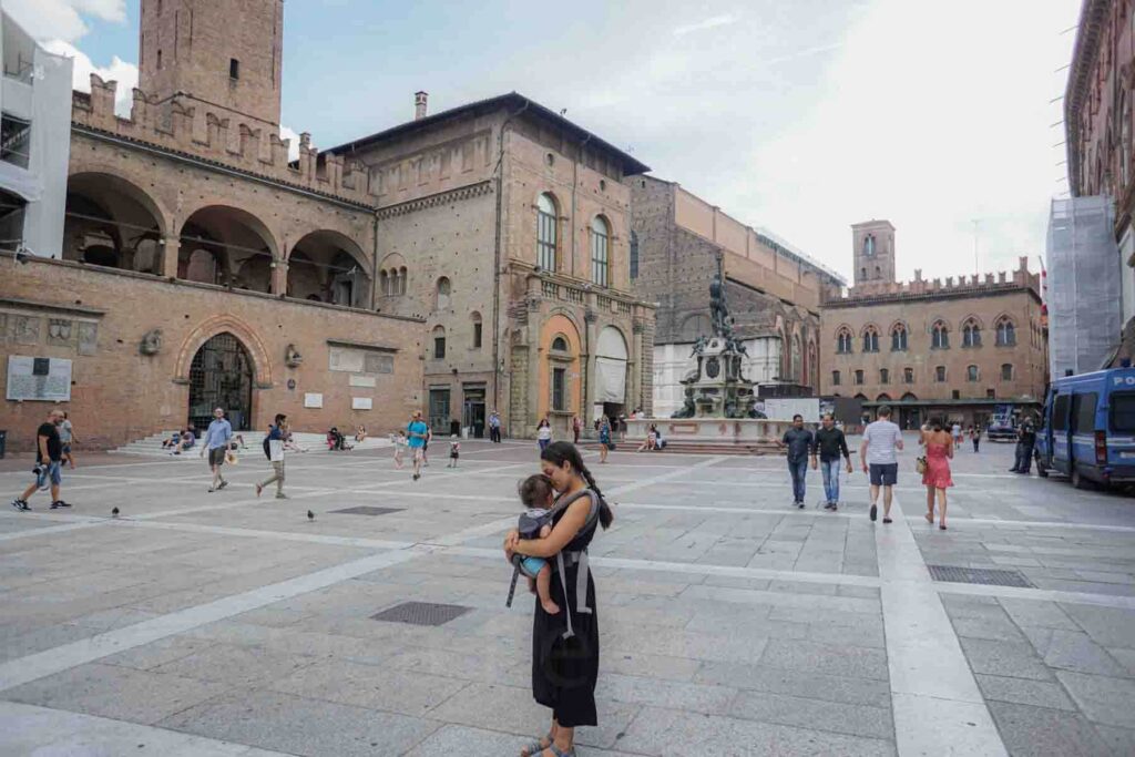 Mother stands in a large historic square in Bologna, Italy holding her baby in an ergo carrier while brick buildings and arches surround them. The baby rests quietly against her chest as people walk through the piazza. Exploring walkable European cities like this can feel manageable even with a baby.