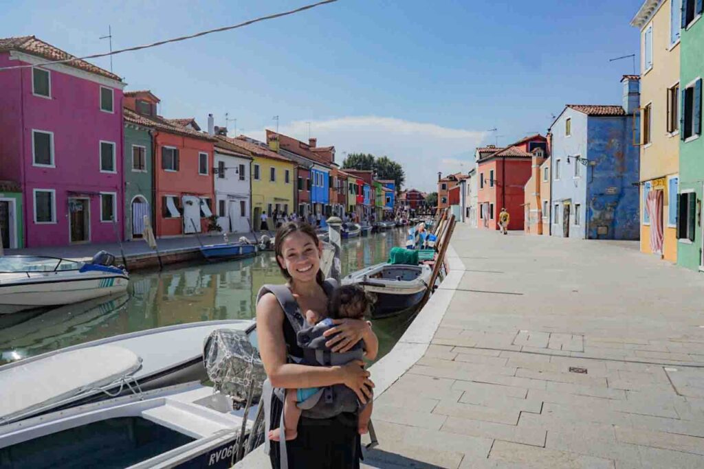 Mother smiles while wearing her baby, content in a carrier along a canal lined with brightly colored houses in Burano, Italy. Small boats float in the water beside the narrow walkway.