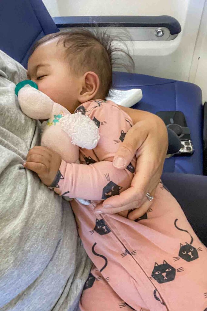 Sleeping baby in pink pajamas cuddles a stuffed pink flamingo wubbanub pacifier while resting in a parent’s arms on an airplane seat. The cabin wall and armrest are visible beside them. Flights can feel smoother at with a younger baby when they are still able to nap comfortably in your arms.