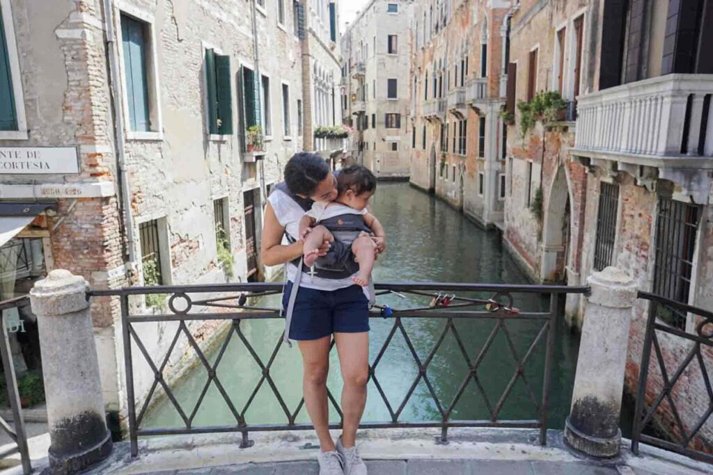 Mother stands on a small bridge over a narrow canal in Venice, Italy while wearing her baby in a front ergobaby carrier. Weathered buildings and green shutters line the waterway behind them. Navigating bridges and cobblestone streets can be easier with a younger baby when babywearing still works well.