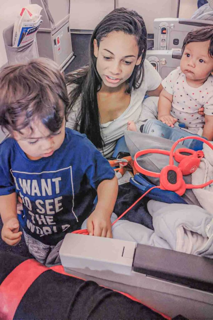 A mom crouches on the floor next to an airplane seat in the bulkhead row with her arm around a baby and her toddler while toys and headphones sit nearby. The toddler’s shirt reads "I WANT TO SEE THE WORLD".