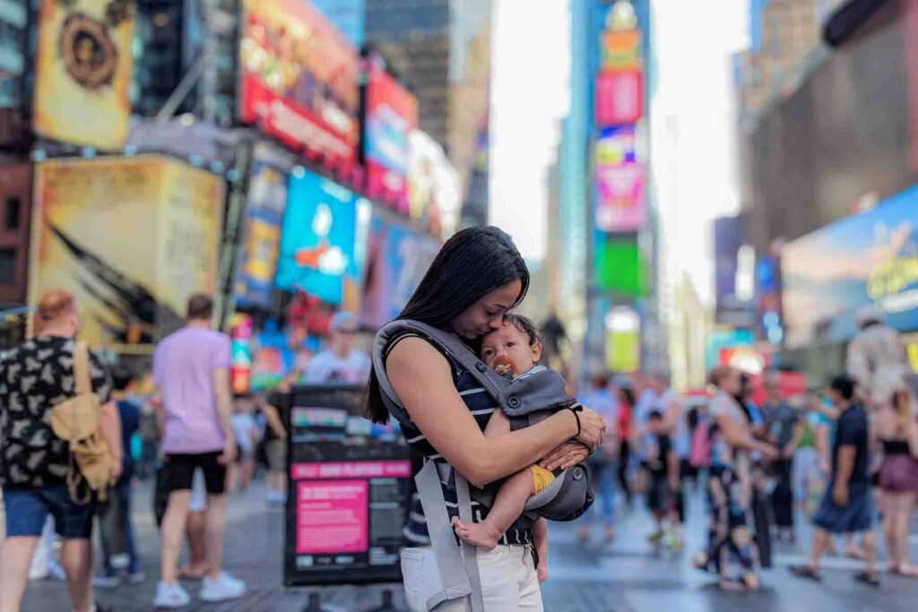 Mother wears her baby in a carrier while standing in the middle of Times Square in New York City among crowds. Colorful digital billboards glow above as people move through the busy street.