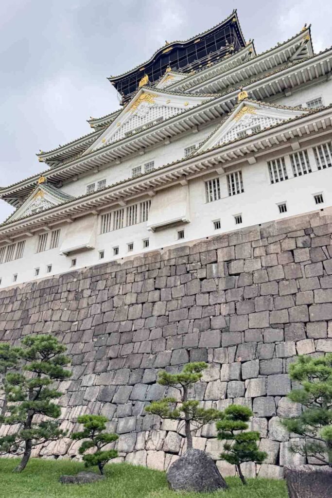 Close view of Osaka Castle rising above massive stone walls with carefully trimmed Bonsai trees at the base. Iconic sights like this are popular to see on first trip to Japan.