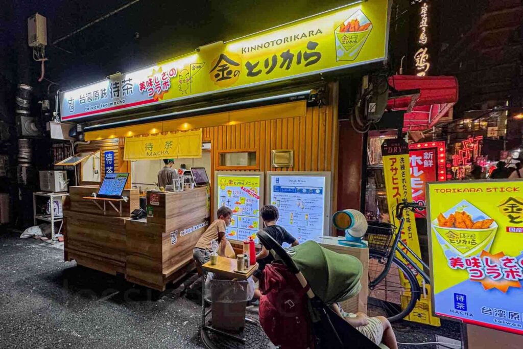Small street food stand in Osaka at night with bright yellow signs for "KINNOTORIKARA" and "MATCHA" while children sit nearby at a tiny table. Food stalls like this offer easy snack options for families.