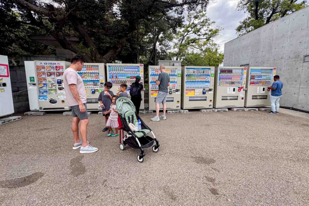 Family with young children eating ice cream standing in front of a row of Japanese vending machines while a stroller sits nearby. Quick snack stops like this are another reason while Japan works so well for families, everything is convenient.
