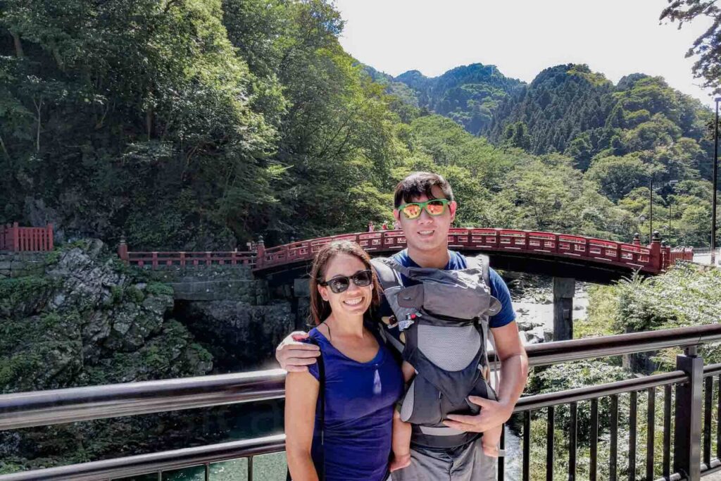 Young parents wearing sunglasses and smiling, while dad has the baby in an Ergo carrier, in front of the Shinkyo Bridge in Nikko on their first Japan trip with a baby.