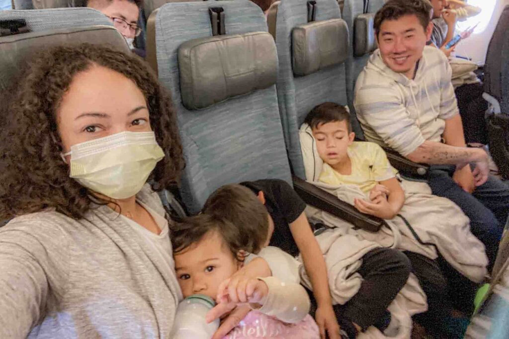 A mom and dad sit airplane seats with three children between them while two children sleep and a baby drinks from a bottle. The scene shows a family managing a long haul flight together.