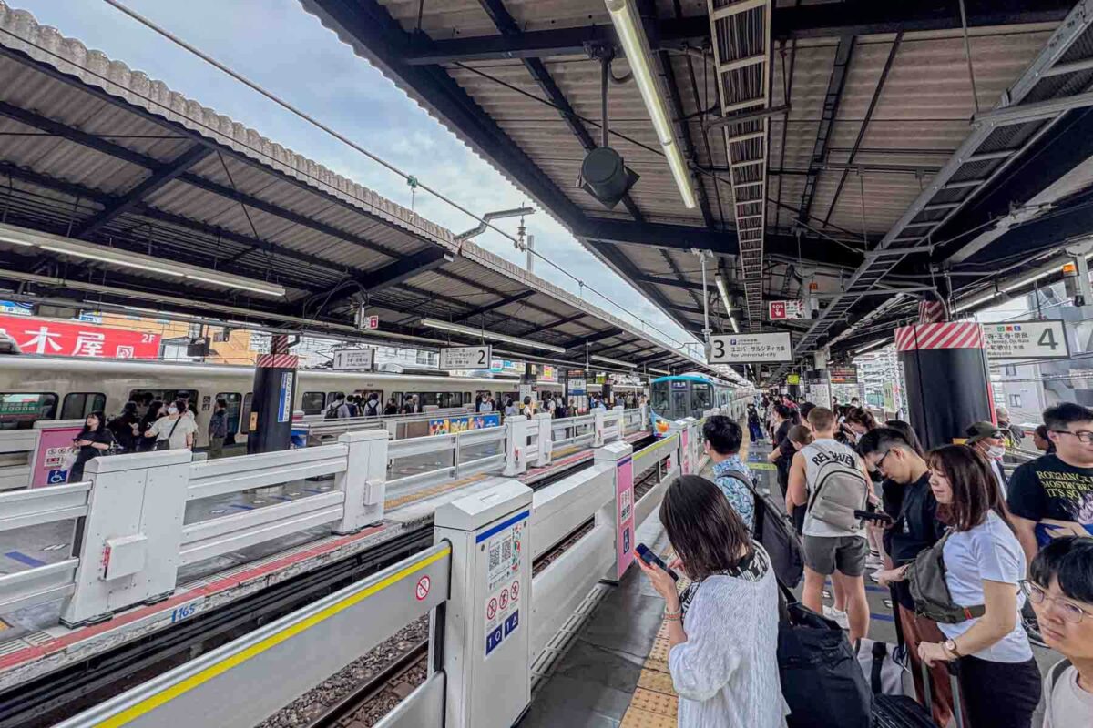 Crowds of passengers line up in organized rows on a busy outdoor train platform in Osaka with clearly marked boarding areas. Demonstrates how structured queues help simplify train travel in Japan with kids.