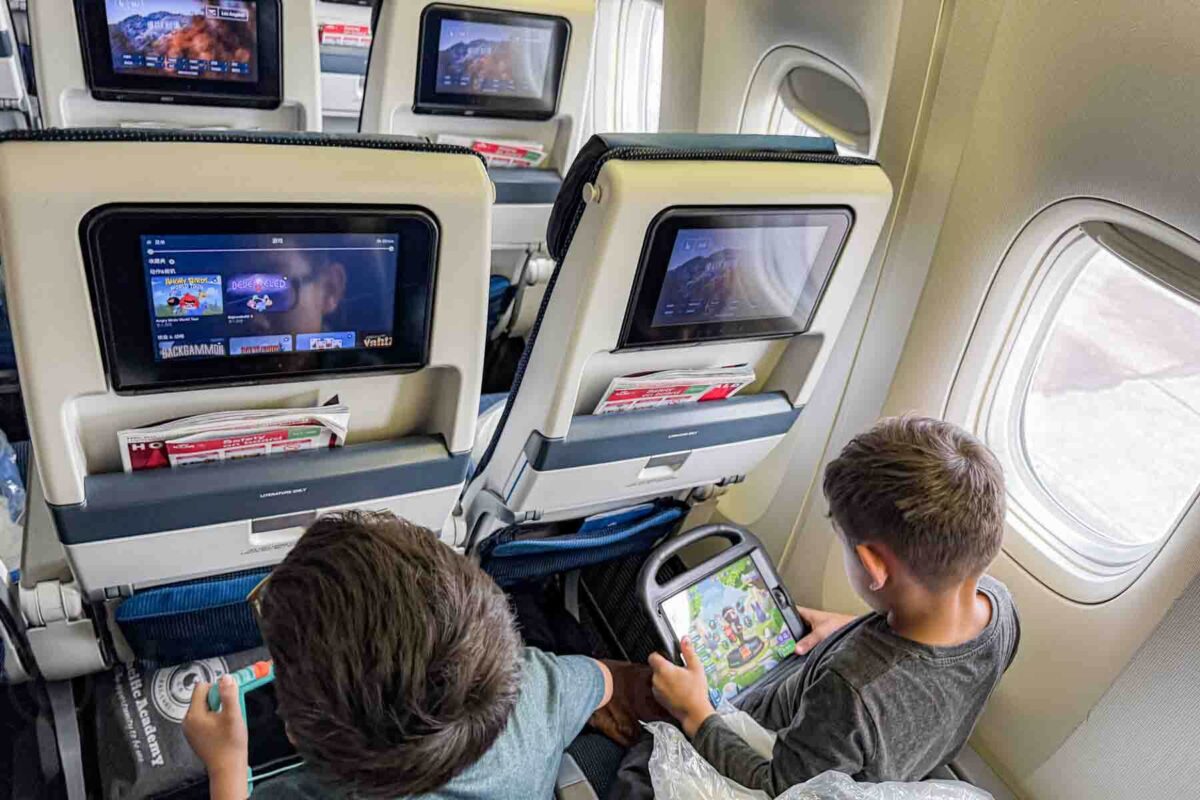 Two elementary-aged boys sit in airplane seats using iPads and the seatback entertainment screens on a long haul flight.