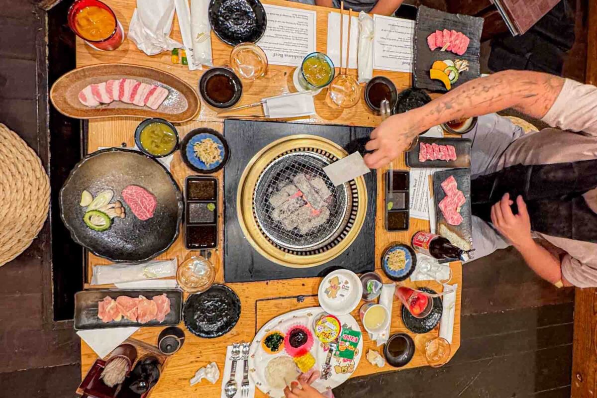 Overhead view of a Japanese barbecue table with a grill in the center in a private dining room at a yakiniku restaurant in Ishigaki as an adult cooks slices of meat while a child sits nearby, surrounded by small dishes, sauces, and drinks. The interactive dining setup shows an easy and engaging way for families to eat together in Japan.
