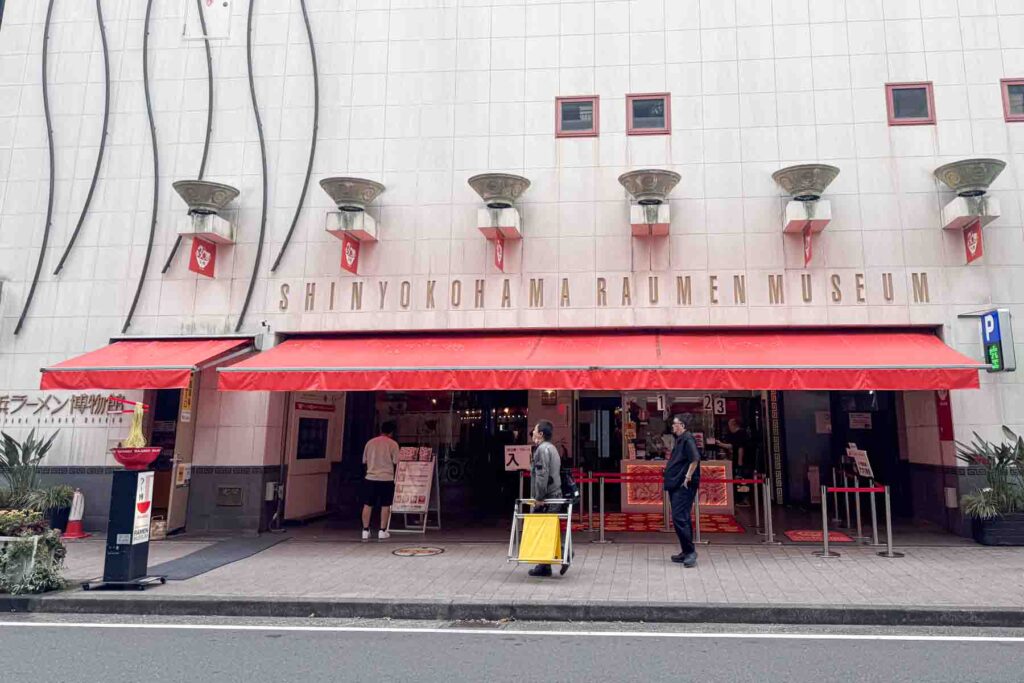 Exterior entrance of the Shin Yokohama Ramen Museum with a red awning and the sign "SHIN YOKOHAMA RAMEN MUSEUM" above the doorway with metal ramen bowls as decor above the sign. Unique attractions like this are fun for families to check out on a trip to Japan.