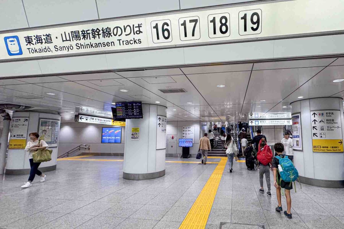 Interior of a busy station with overhead sign reading "Tokaido, Sanyo Shinkansen Tracks 16 17 18 19" as travelers walk toward platforms. Clear signage helps families navigate around Japan with kids by train.