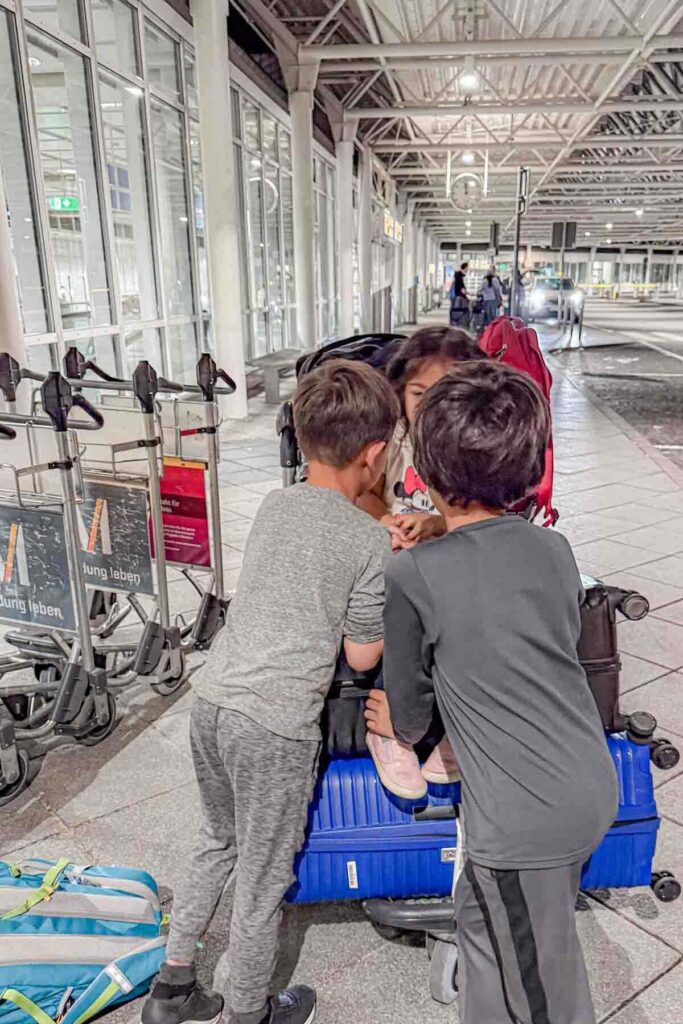 Three siblings gather around a luggage cart stacked with suitcases outside Munich airport near baggage trolleys. The children wait together after a long international flight.