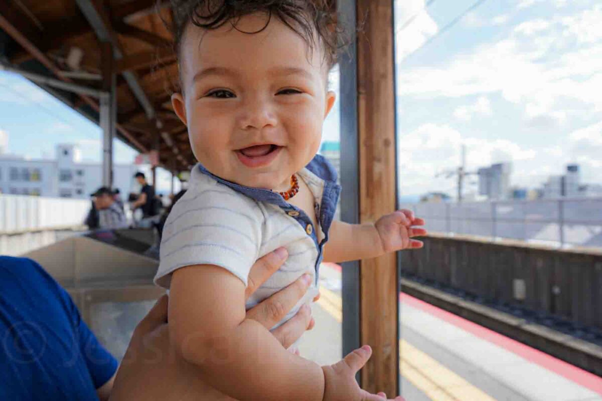 Smiling baby held up near an outdoor train platform with city buildings in the background. Captures a joyful moment while traveling through Japan with a baby.
